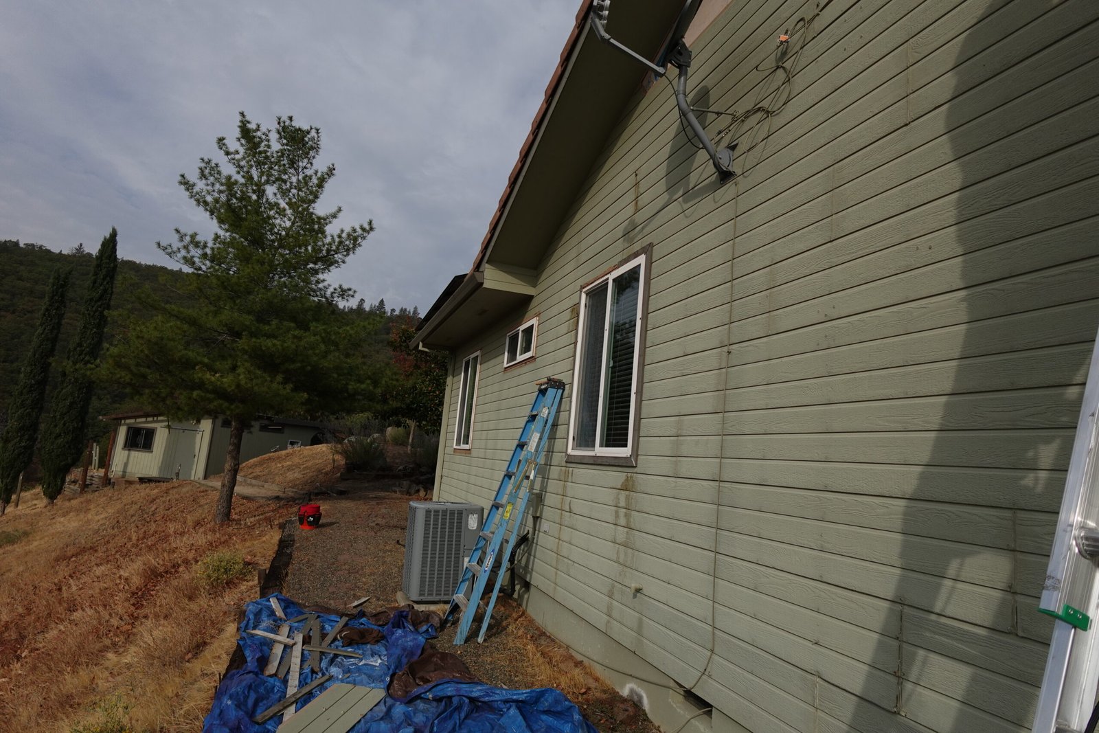 “Side exterior of a residential home with green horizontal siding, a ladder leaning against the wall, tarps and tools on the ground, and a hillside landscape with trees in the background.