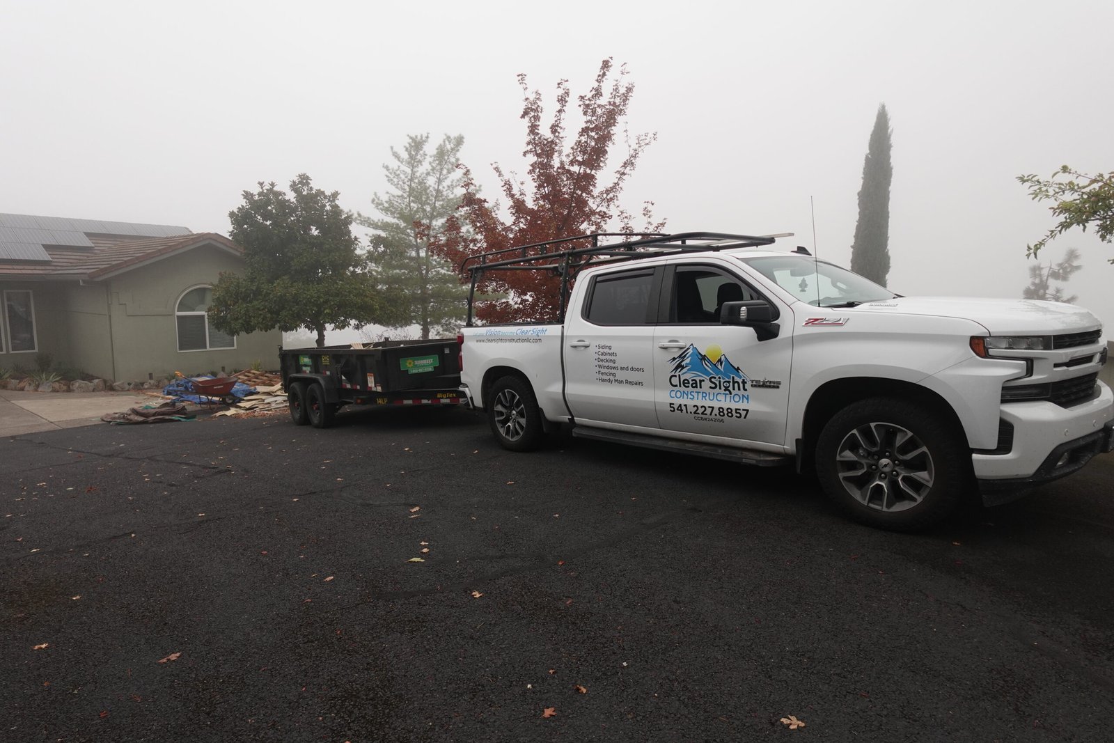 White Clear Sight Construction pickup truck with company logo towing a flatbed trailer, parked on a foggy residential driveway with trees and construction debris visible.